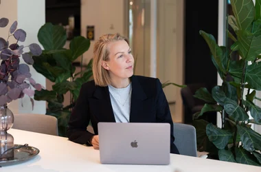 Kvinna i kavaj som sitter framför dator i kontorsmiljö och tittar upp till höger.